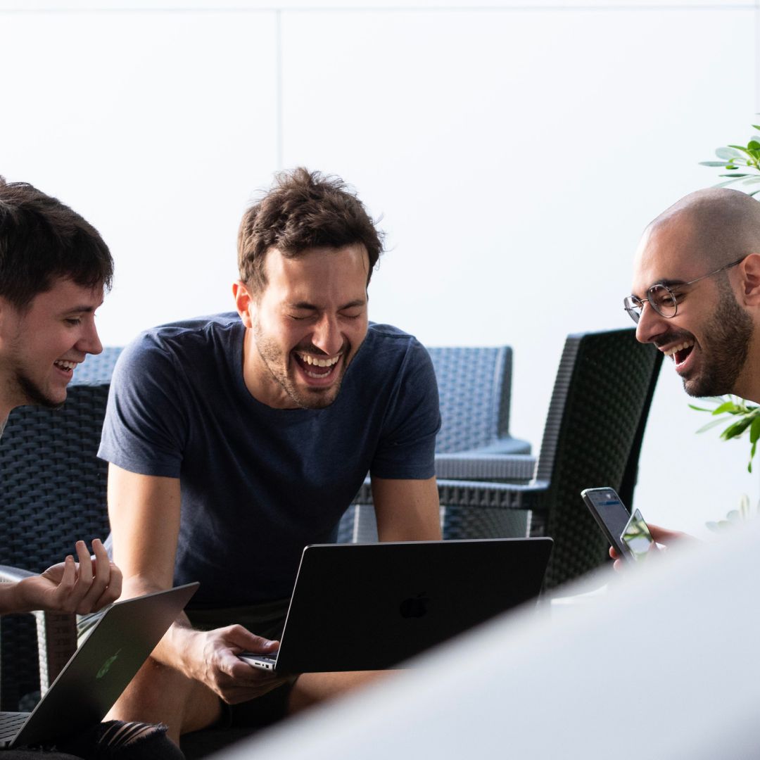 3 men looking at a laptop. The middle one is laughing.