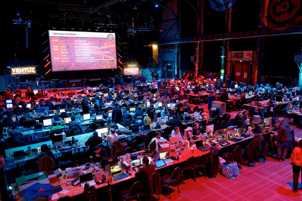 Revision party, the party hall filled with computers on desks. The main screen is showing the timetable in the background. Image credit Viscid