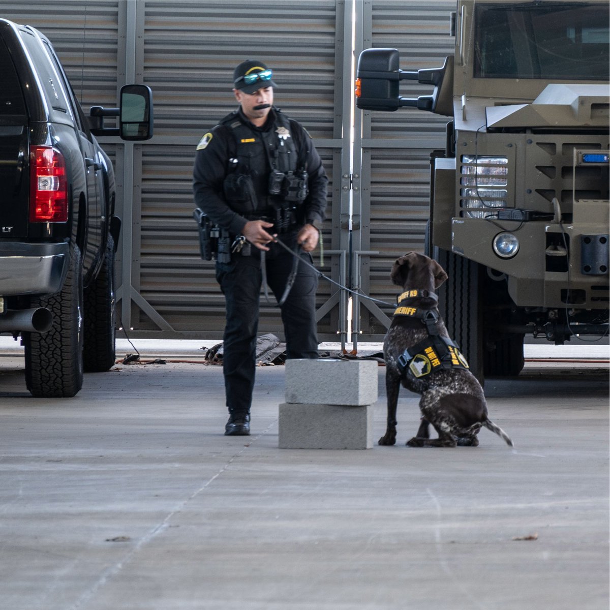 Police officer in tactical gear holding a leash attached to a sitting K9 unit inside a garage with vehicles.